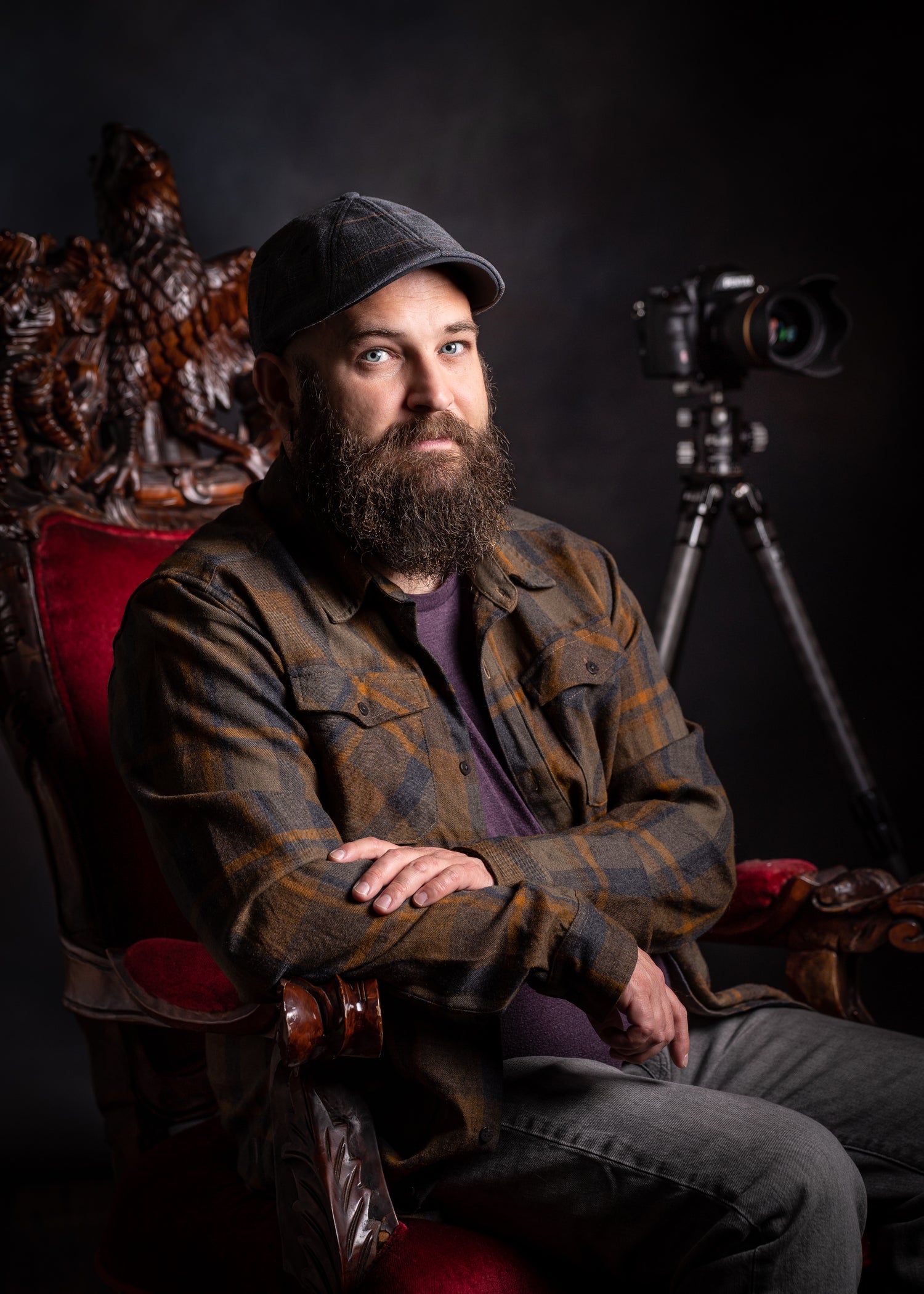 Man with a beard wearing a plaid jacket and cap sitting in front of a camera on a tripod.