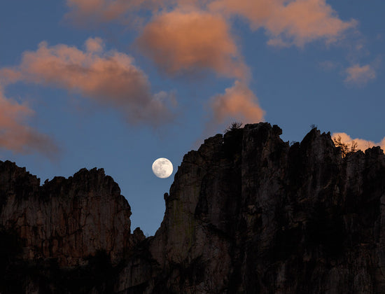 The full moon rises through the gunsight notch silhouetted against a blue sky at Seneca Rocks in West Virginia with twilight pink clouds.